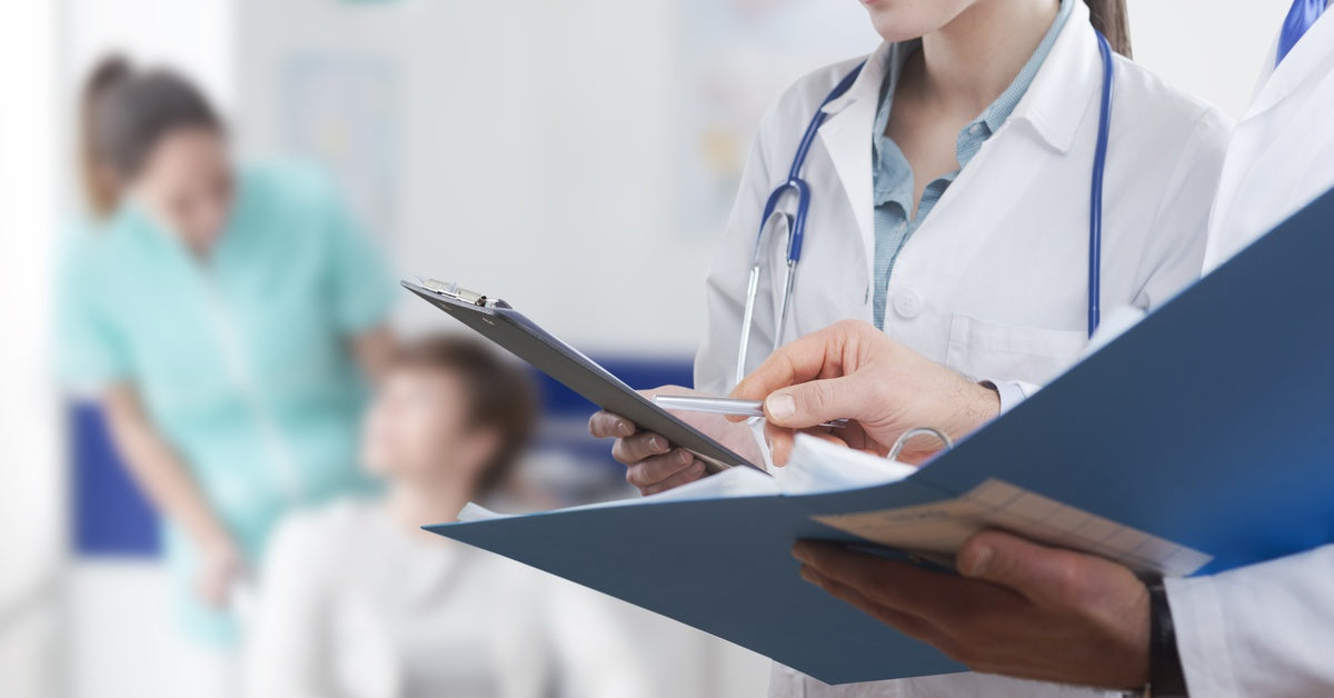 A person in a white coat is standing next to another person who is out of frame. There are several people blurry in the background