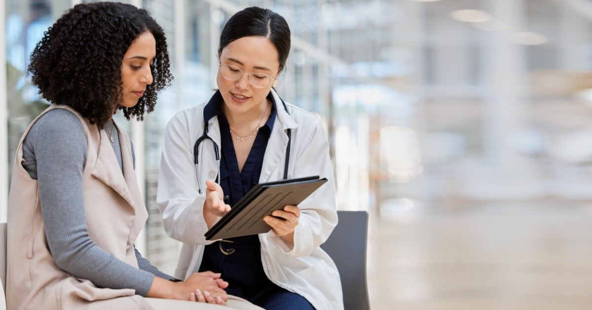 A female doctor is sitting next to a woman on a bench. The doctor is holding a tablet and gesturing to it with her hand.