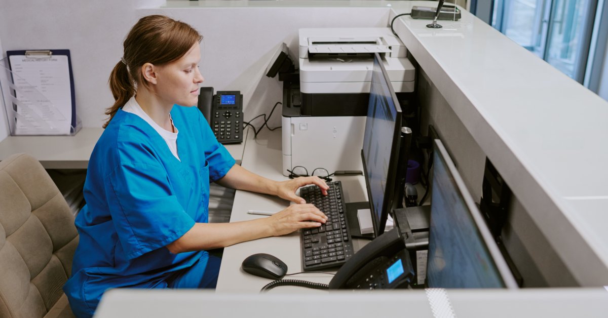 A woman in blue scrubs is sitting at a desk and typing on a computer. There is a phone next to her on a desk.