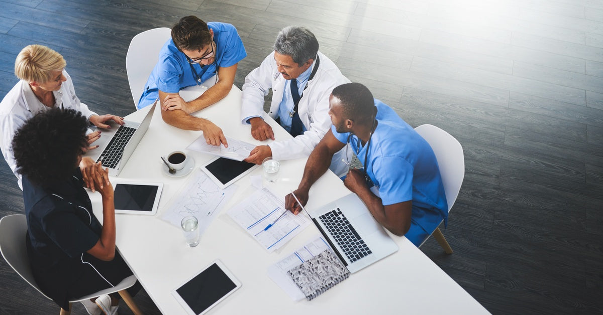 A group of people are sitting at an oval-shaped table. They are all wearing medical gear, such as scrubs and white coats.