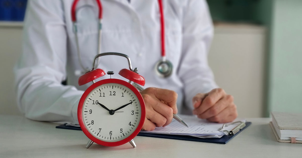 A red alarm clock sitting on the desk in front of a person wearing a stethoscope, writing on papers on a clipboard.