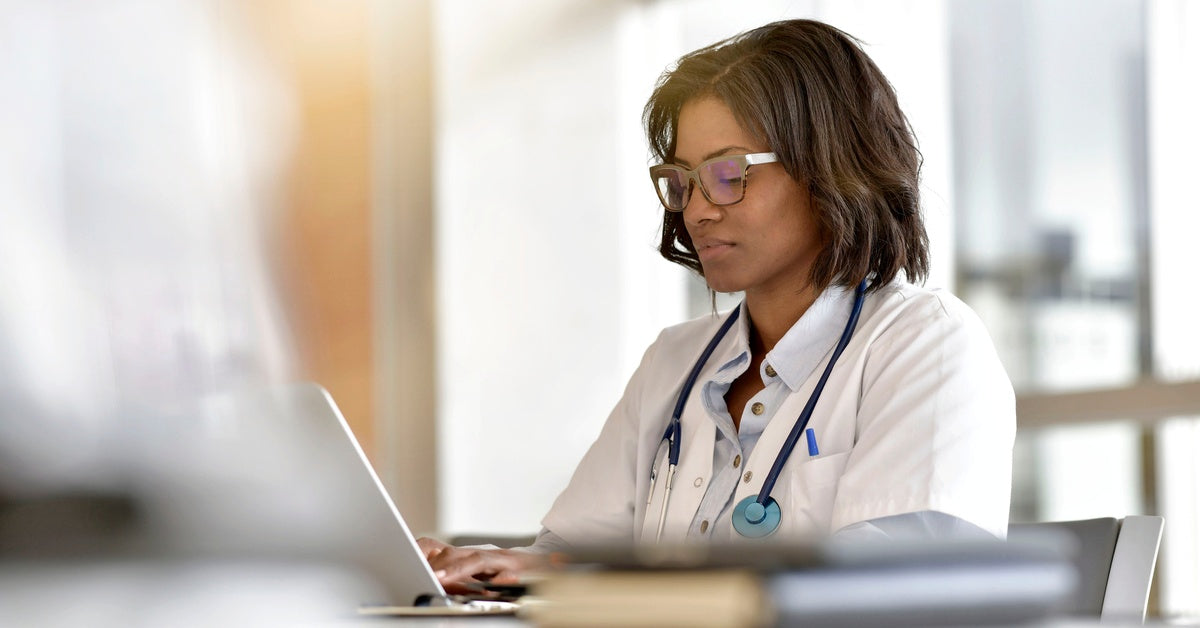 A Black female doctor in a white coat with a stethoscope around her neck sitting and typing on a laptop on a desk.