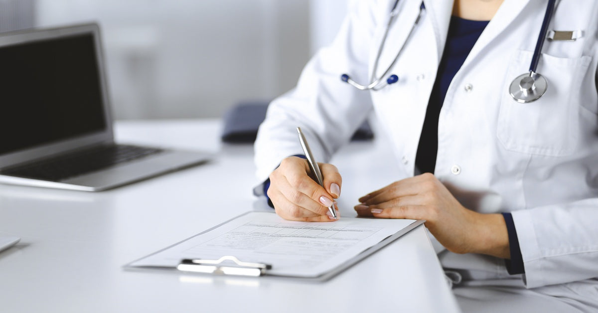 A woman wearing a lab coat and stethoscope, using a pen to write on papers attached to a clipboard sitting on a white table.