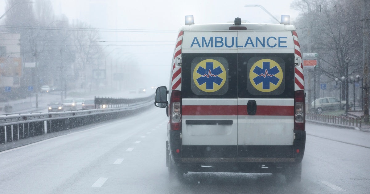An ambulance driving along an empty, triple-lane road during an overcast and wet, snowy day. Its siren lights are off.