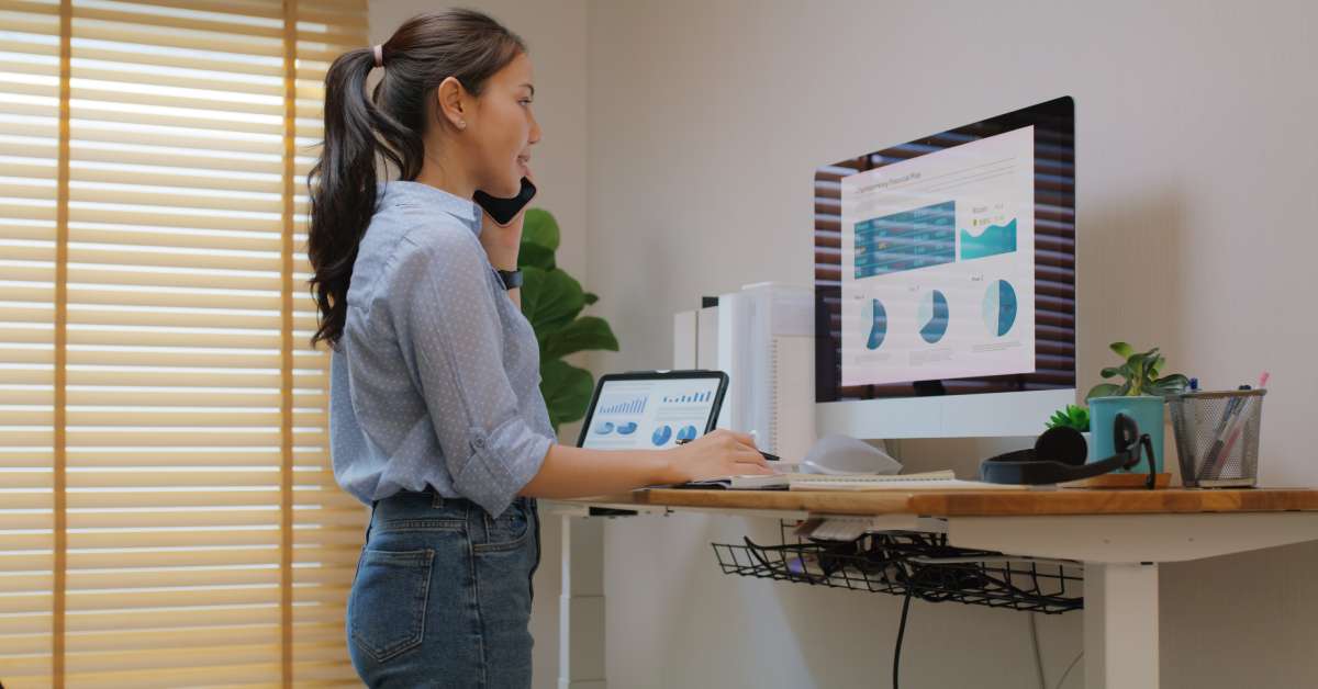 A close-up of a young woman on the phone while working on her desktop computer and tablet at a standing desk.