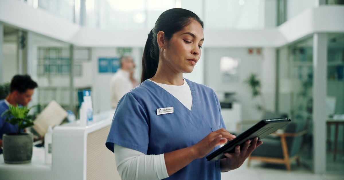 A woman wearing blue scrubs looks at a tablet in her hand. She is in a room with several other people.