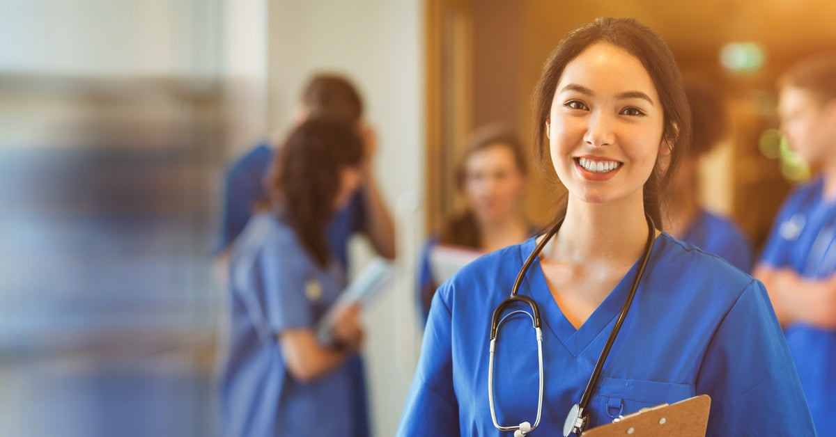 A woman in blue scrubs wears a stethoscope around her neck. She’s smiling and there is a group of people behind her.