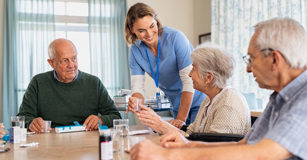 A woman in blue scrubs is standing next to a table. At the table are several older individuals sitting