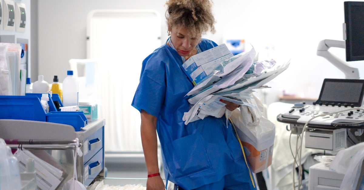 A woman in blue scrubs is leaning over to pick up some materials that are left in a drawer.