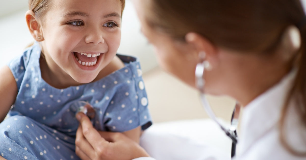 A young child is smiling as a doctor in a white coat listens to her heartbeat and lungs with a stethoscope.