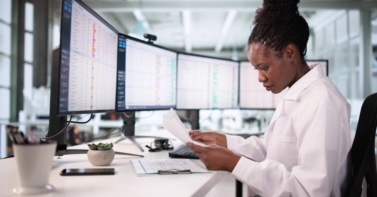 A woman sits at a desk and is looking at a paper she is holding. There are several computer monitors.