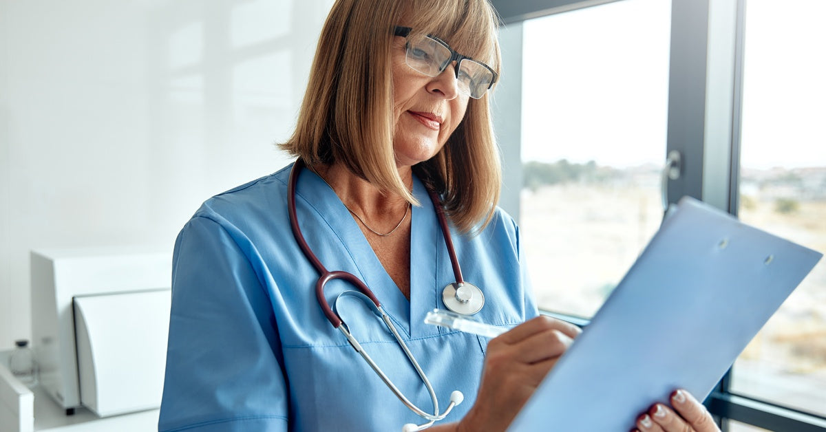 A woman in a medical office wears scrubs and a stethoscope, holds a clipboard, and writes on it with a pen.