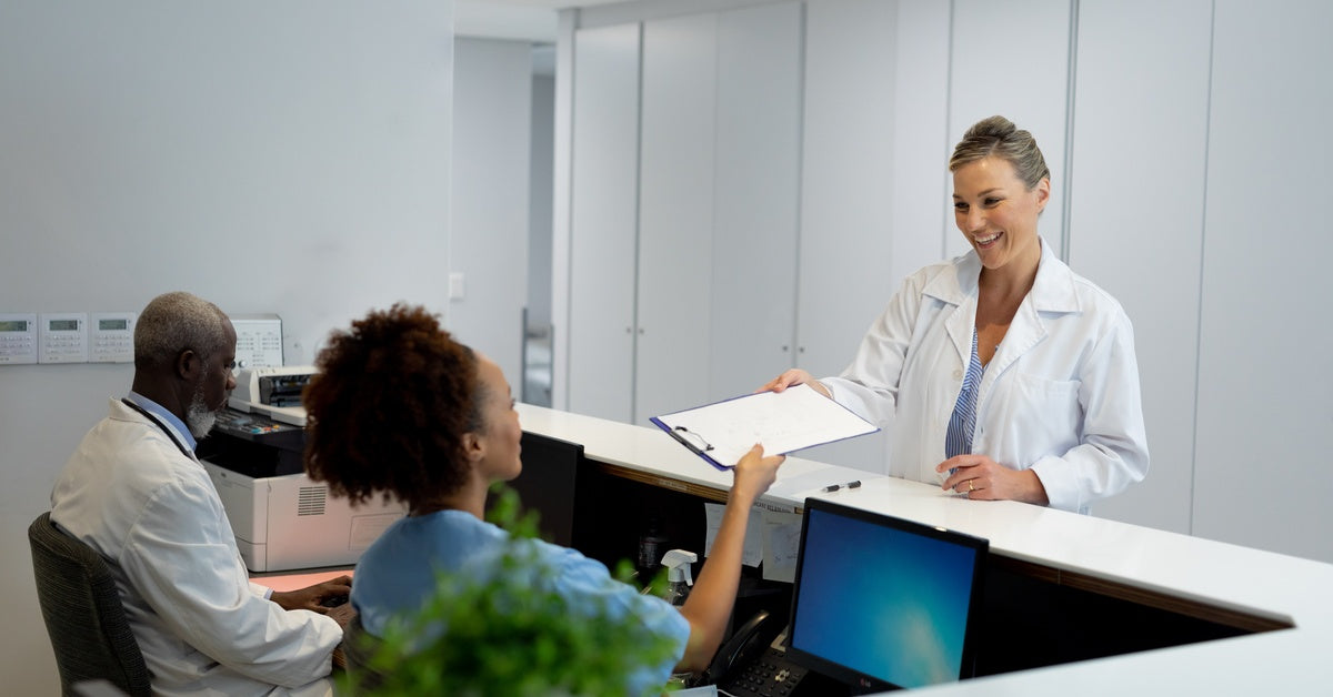 A clipboard with papers on it passes between a woman in a medical coat and a woman sitting behind a desk.