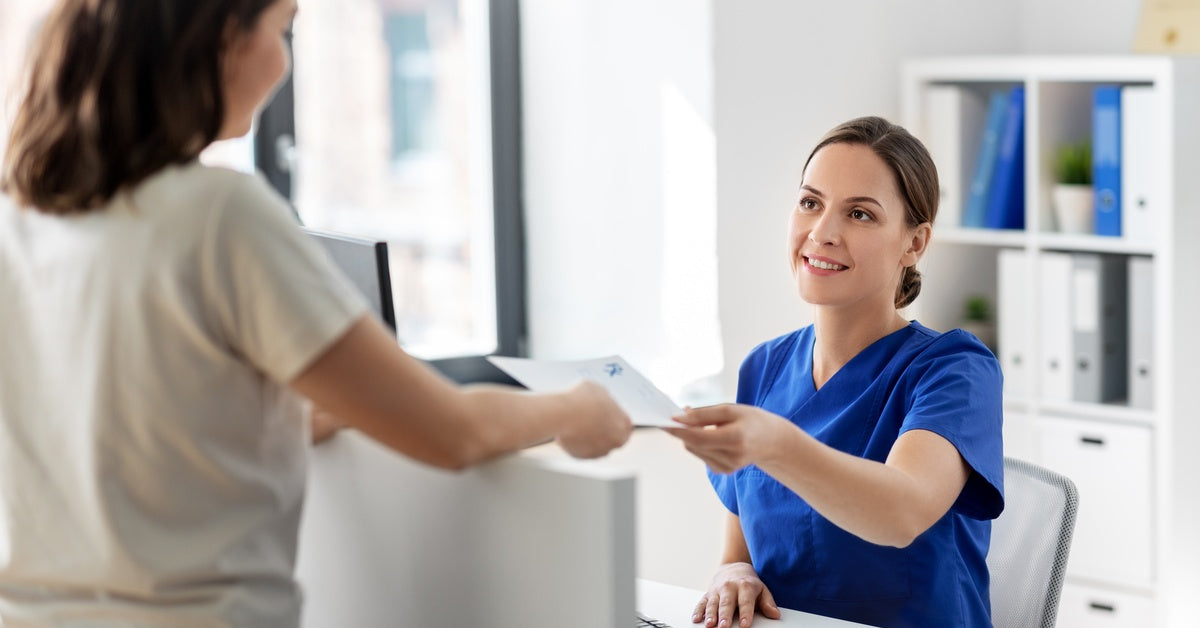 A smiling woman in blue scrubs sitting behind a desk in a health care environment accepting a paper from a coworker.