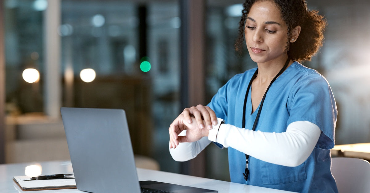 A woman in blue scrubs looks at the watch on her wrist while sitting in front of a laptop at work at night.