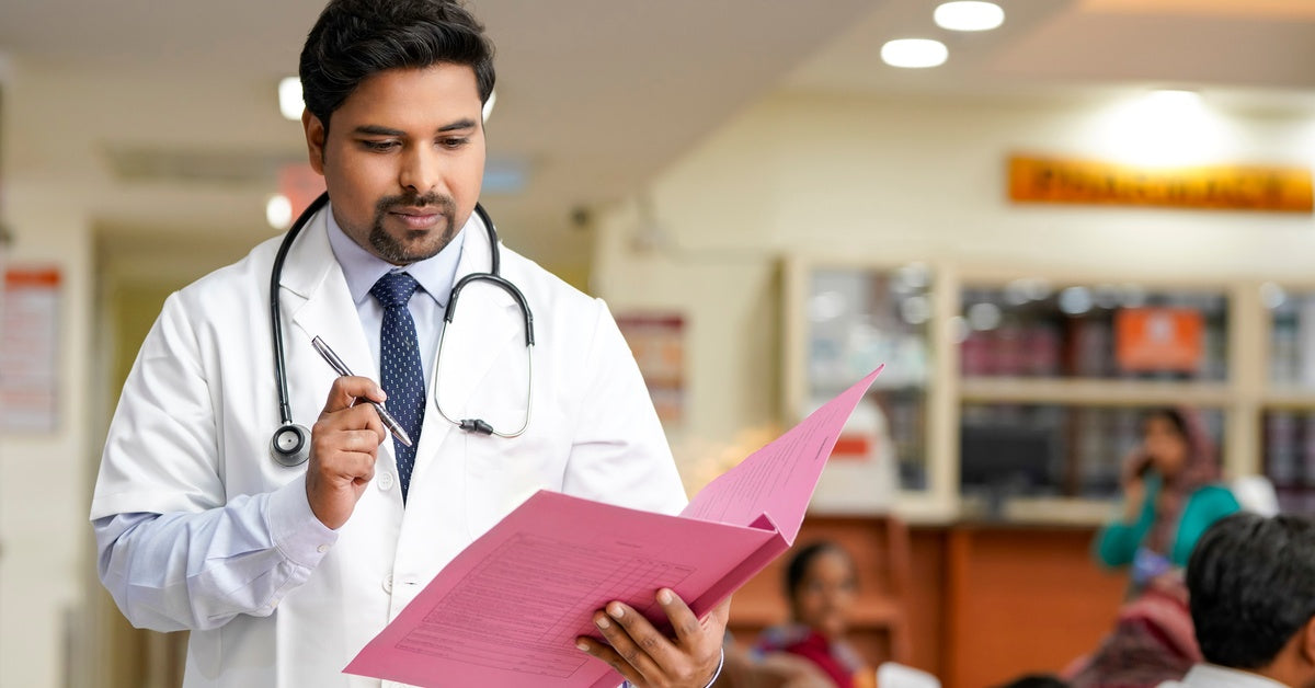 A man in a white doctor's coat with a stethoscope on him holds a pink folder and a pen inside a healthcare setting.
